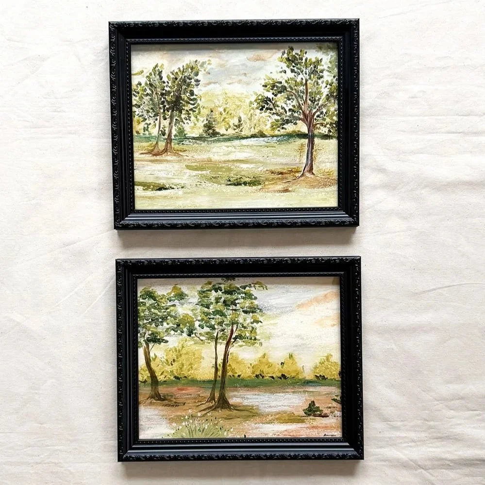 Two framed landscape paintings of trees by a river, displayed vertically on a textured off-white fabric background. The frames are black with ornate detailing.