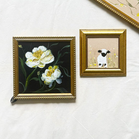 Two framed wall art pieces displayed on a light fabric surface. The larger square frame on the left contains a dark painting of white flowers with green stems and yellow centers, set in a gold ornate frame.