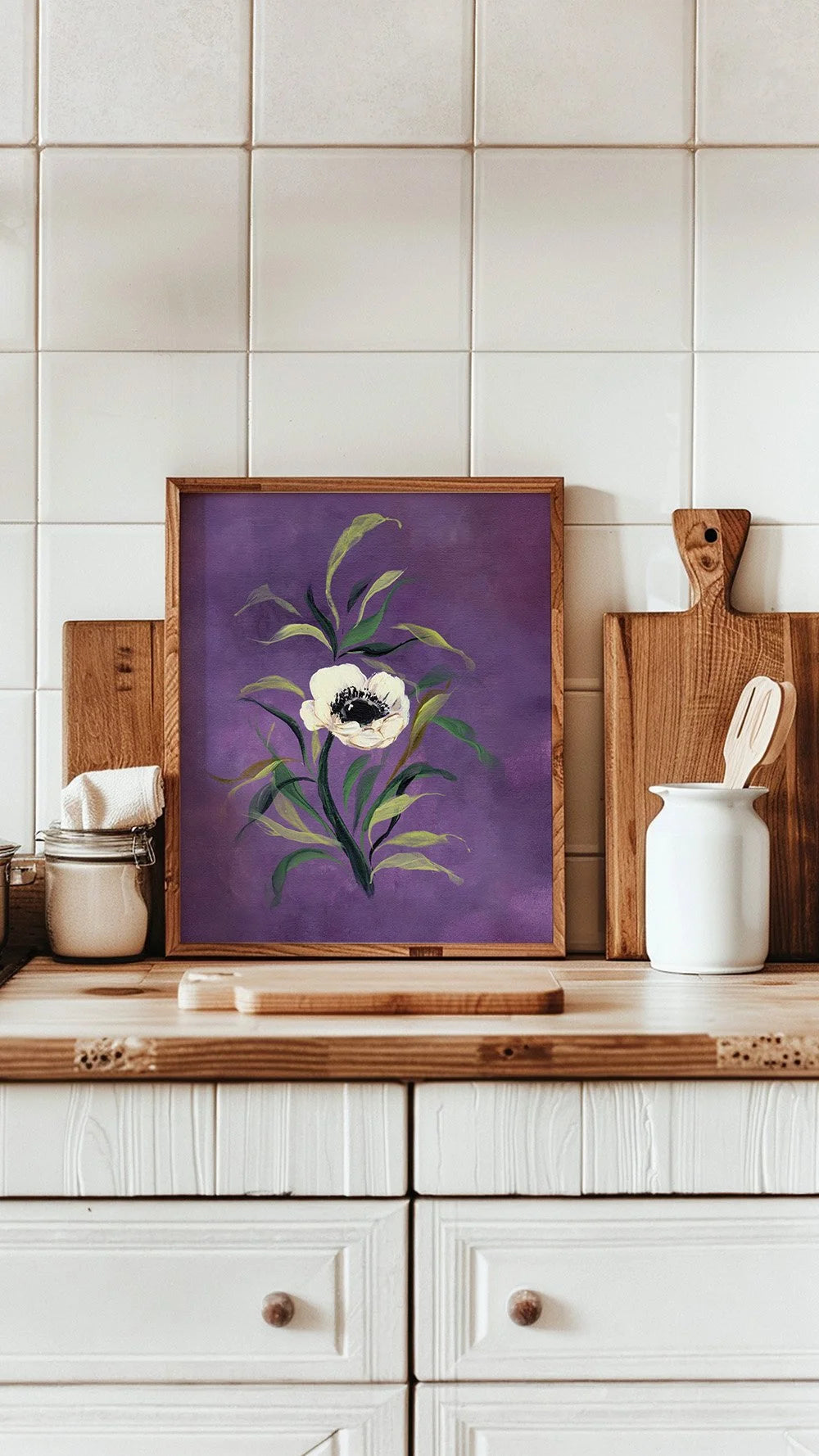 A framed botanical art print featuring a white anemone flower on a purple background, styled on a wooden kitchen counter against a white tiled wall.