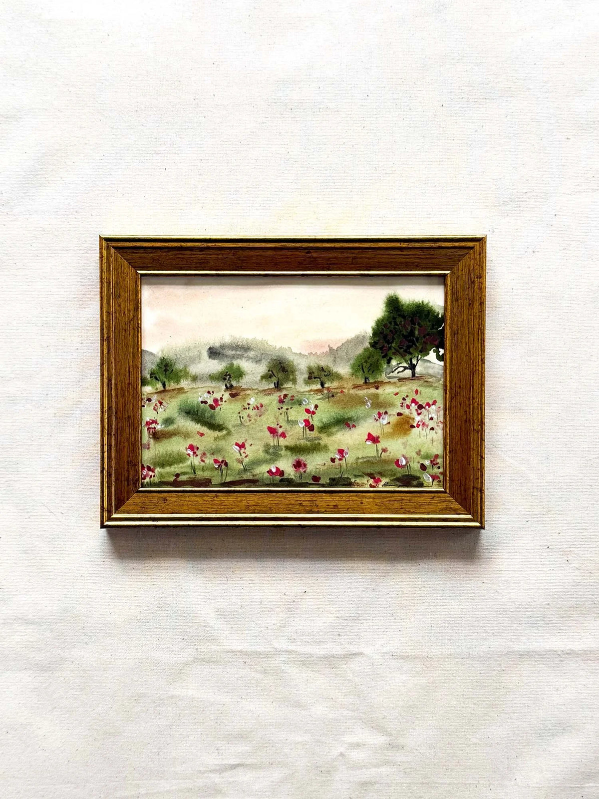 A small framed landscape painting featuring a field of pink and white flowers, green trees, and distant hills, housed in a brown wooden frame and displayed against a textured off-white background.