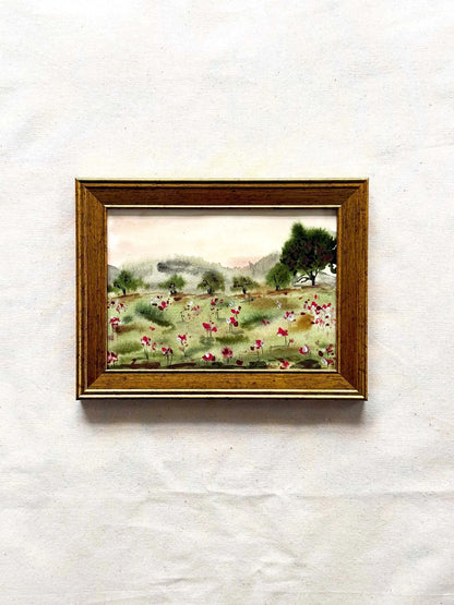 A small framed landscape painting featuring a field of pink and white flowers, green trees, and distant hills, housed in a brown wooden frame and displayed against a textured off-white background.