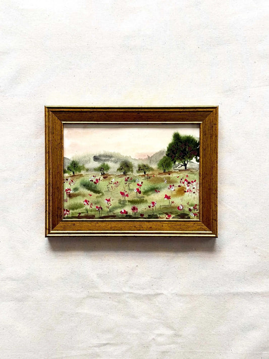 A small framed landscape painting featuring a field of pink and white flowers, green trees, and distant hills, housed in a brown wooden frame and displayed against a textured off-white background.