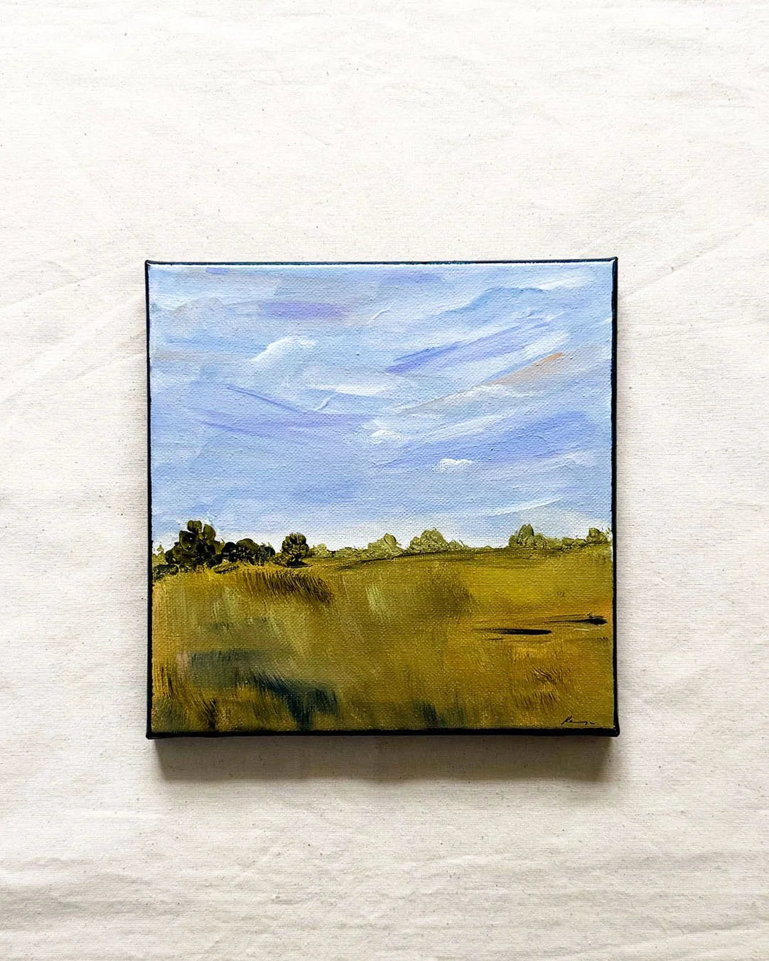A canvas painting of a landscape, featuring a golden-yellow field under a blue sky with white clouds, displayed on a textured off-white fabric background.