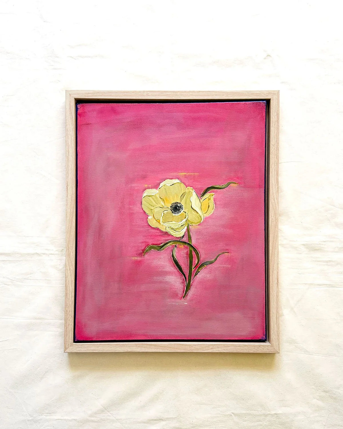 A framed painting of a single yellow flower with green stems on a textured pink background, displayed on a white fabric backdrop.