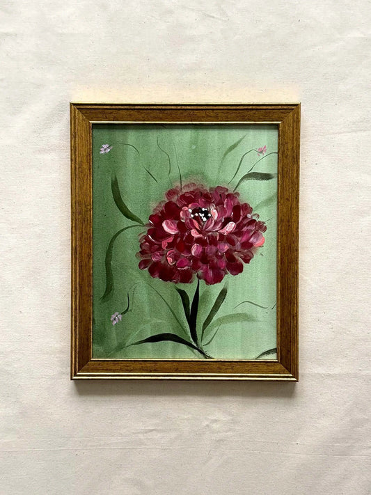 A framed painting of a burgundy peony flower with green leaves on a light green background, housed in a gold-toned wooden frame and displayed on a textured off-white wall.
