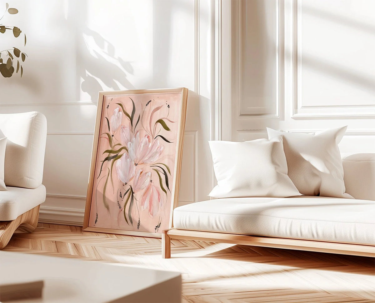 A framed floral painting with soft pink flowers and green leaves leaning against a wall in a bright, modern living room with white furniture and wood flooring.