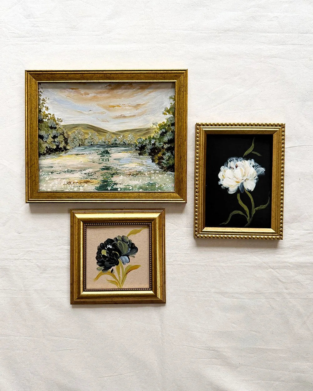 Three framed botanical and landscape paintings with ornate gold frames hanging on a textured white wall.