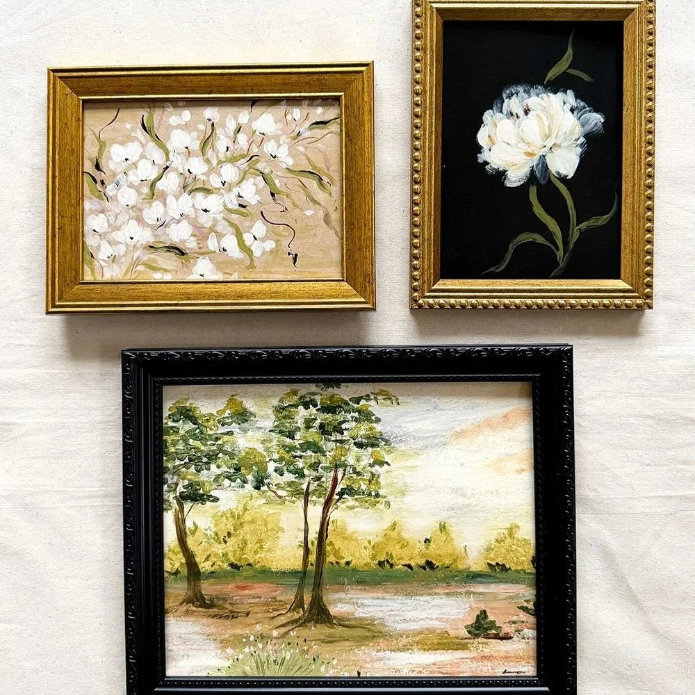 Three framed paintings displayed on a white fabric background: a gold-framed floral artwork at upper left, a gold-framed black-and-white flower painting at upper right, and a black-framed landscape painting at lower center.