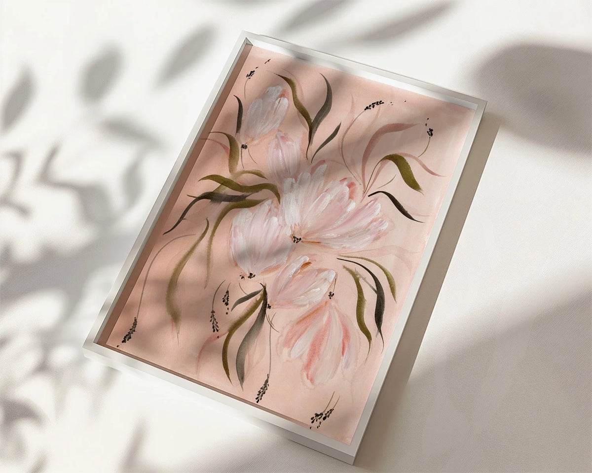A framed floral art print featuring soft pink tulips with green leaves on a pale pink background, shown at an angle on a white surface with dappled shadows.