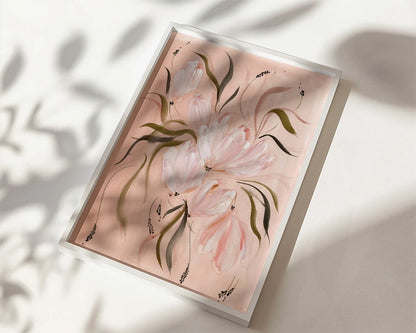 A framed floral art print featuring soft pink tulips with green leaves on a pale pink background, shown at an angle on a white surface with dappled shadows.