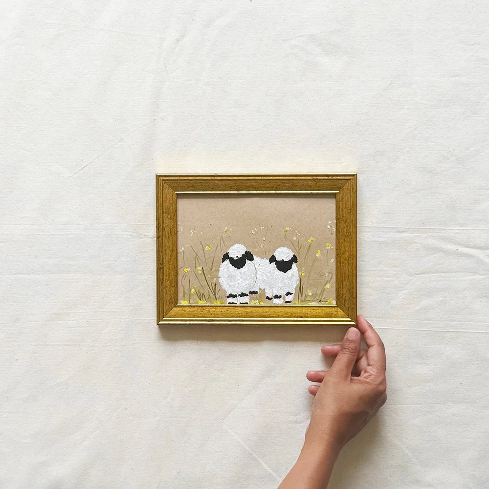 A small framed artwork showing two white sheep with black faces standing in a field of yellow flowers, held by a person's hand against a light fabric background.