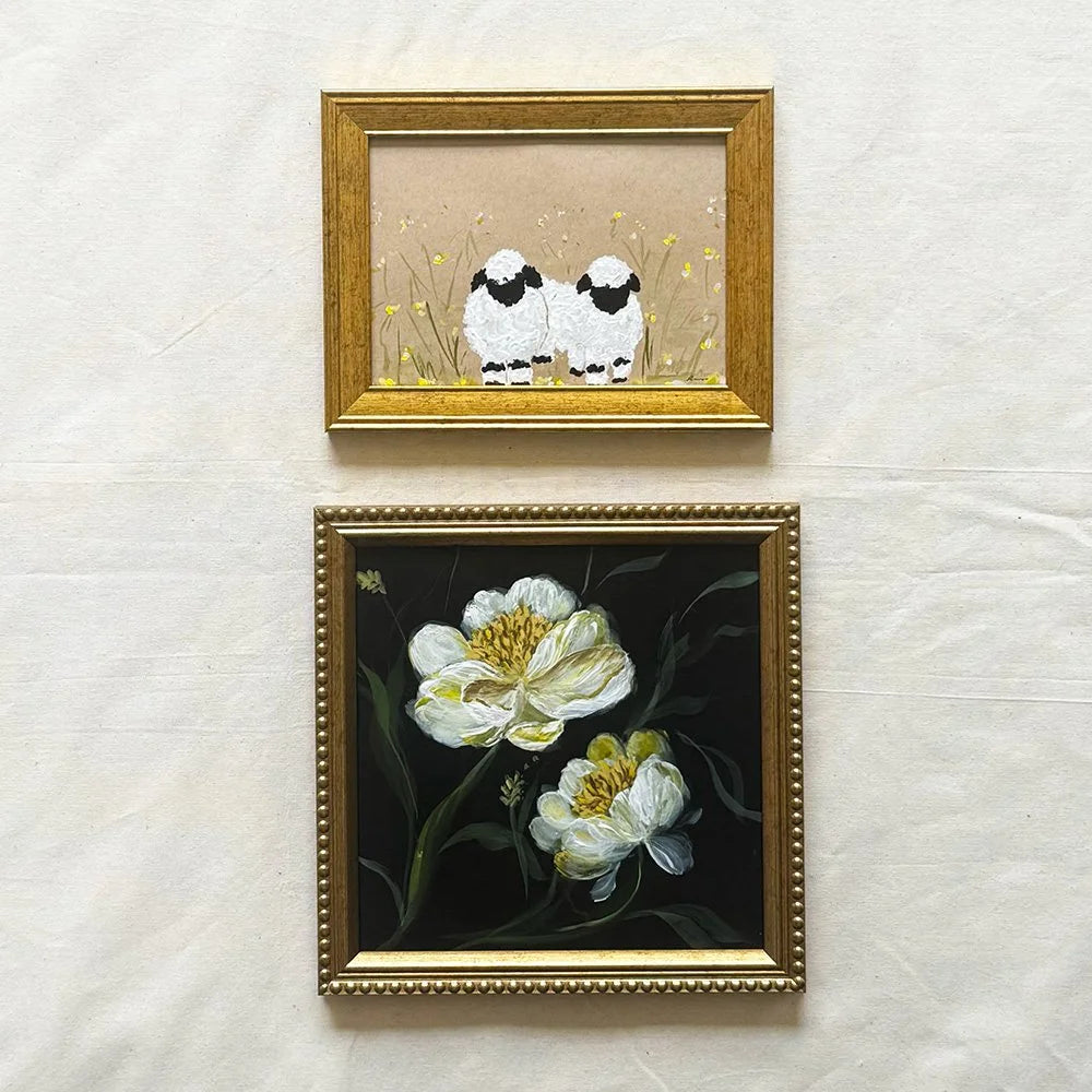 Two framed artworks hanging on a light fabric background. The top frame shows two small white sheep with black faces in a beige field, and the bottom frame shows white flowers with yellow centers on a dark background, both in ornate gold frames.
