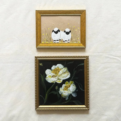 Two framed artworks hanging on a light fabric background. The top frame shows two small white sheep with black faces in a beige field, and the bottom frame shows white flowers with yellow centers on a dark background, both in ornate gold frames.