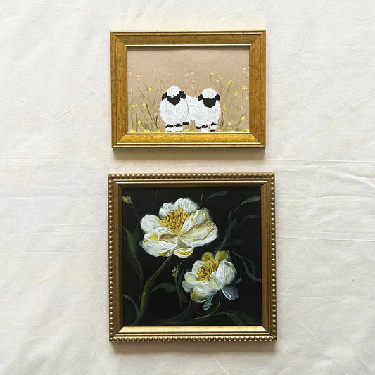 Two framed artworks hanging on a light fabric background. The top frame shows two small white sheep with black faces in a beige field, and the bottom frame shows white flowers with yellow centers on a dark background, both in ornate gold frames.