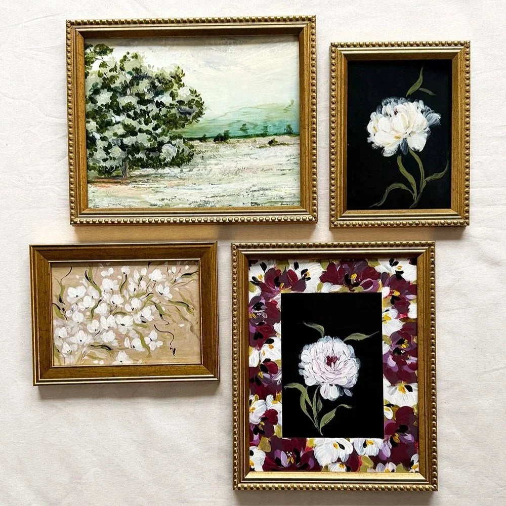 Four framed floral and landscape paintings arranged on a light textured wall. The frames are ornate gold-toned, and the paintings feature white flowers, a green tree by the sea, and a central white flower surrounded by dark purple and burgundy floral details.