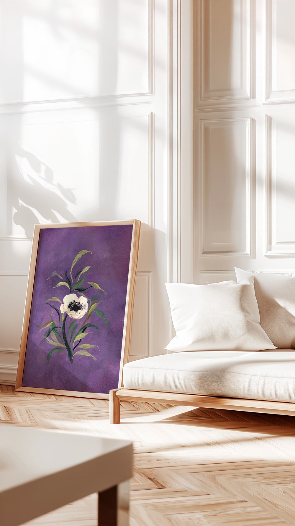 A framed art print featuring a purple background with a white flower and green leaves, leaning against a white paneled wall in a bright, modern living room next to a white sofa.