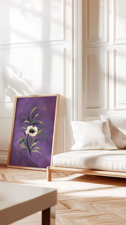 A framed art print featuring a purple background with a white flower and green leaves, leaning against a white paneled wall in a bright, modern living room next to a white sofa.