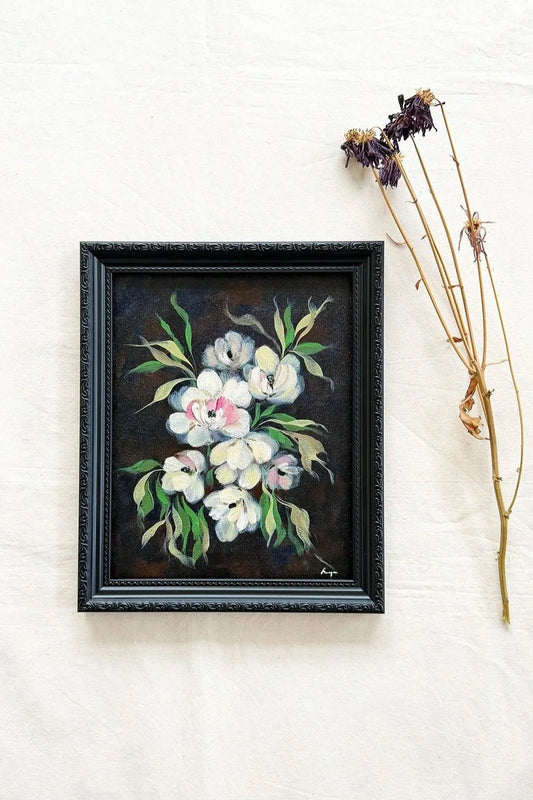A framed floral painting featuring white and pink flowers with green leaves on a dark background, set in a black ornate frame and styled on a white fabric surface next to dried stems.