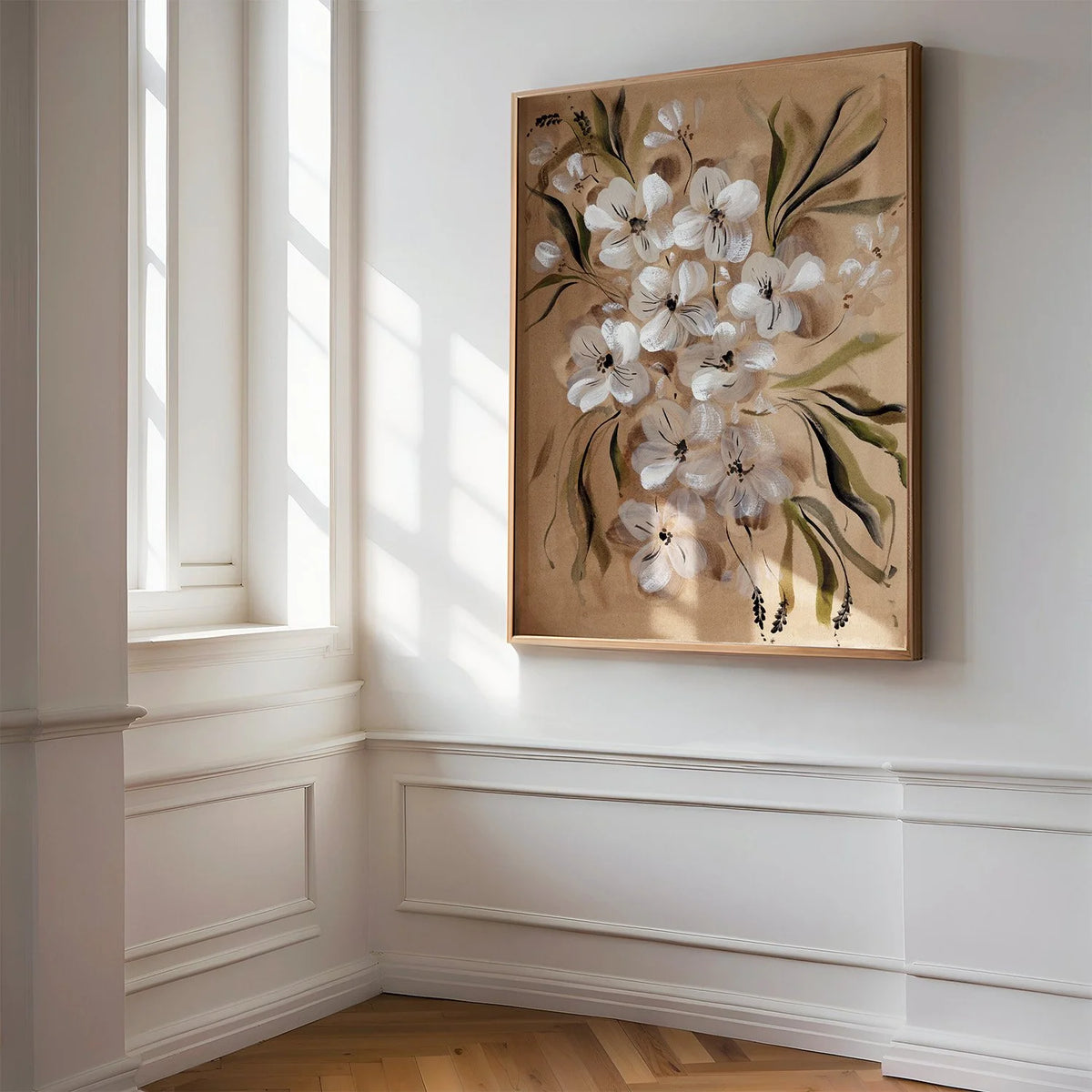 A framed floral painting featuring white blossoms and green leaves on a warm beige background, displayed on a white wall in a sunlit room with wood flooring and white wainscoting.
