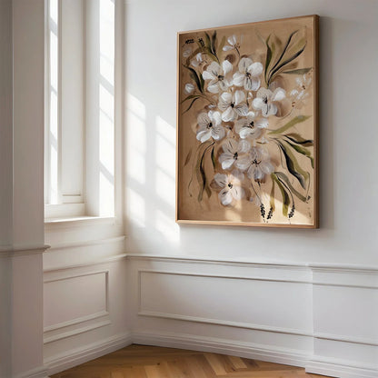 A framed floral painting featuring white blossoms and green leaves on a warm beige background, displayed on a white wall in a sunlit room with wood flooring and white wainscoting.