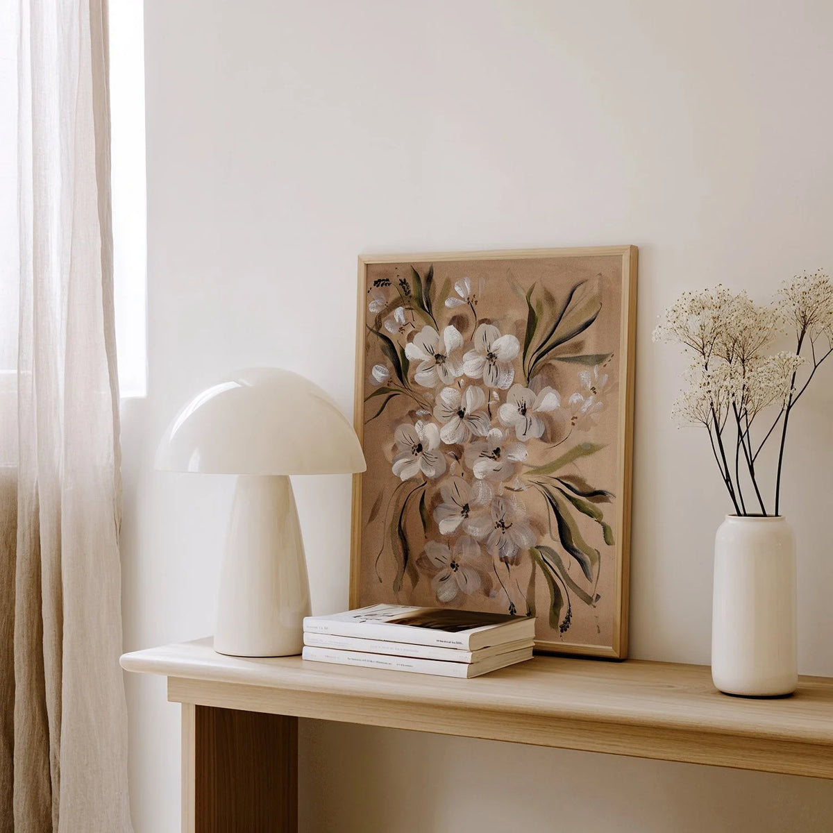 A framed floral art print with white blossoms and green leaves leaning on a light wood console table, styled with a white mushroom lamp, stacked books, and a white vase with dried flowers in a bright room.