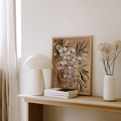A framed floral art print with white blossoms and green leaves leaning on a light wood console table, styled with a white mushroom lamp, stacked books, and a white vase with dried flowers in a bright room.