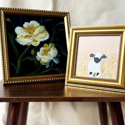 Two framed art pieces displayed on a wooden table against a light fabric backdrop. The larger left frame shows a dark painting of white and yellow flowers with green stems, and the smaller right frame shows a small white lamb with a black face in a field of yellow flowers on a pale pink background.
