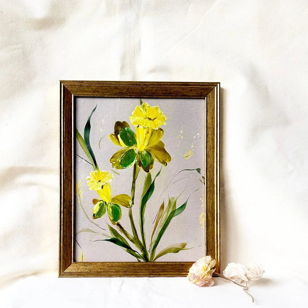A framed painting of yellow flowers with green leaves on a light gray background, set in a rustic brown wooden frame and styled on a white fabric backdrop with dried flowers.