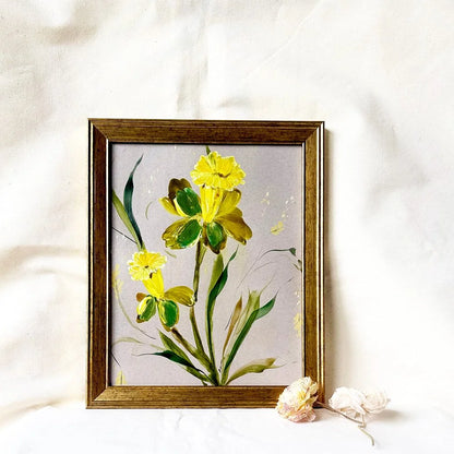 A framed painting of yellow flowers with green leaves on a light gray background, set in a rustic brown wooden frame and styled on a white fabric backdrop with dried flowers.