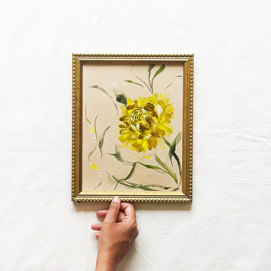 A framed painting of a yellow flower with green leaves on a textured beige background, held by a hand against a white fabric backdrop.