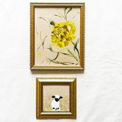 Two framed wall art pieces hanging on a white wall. The larger top frame shows a yellow flower with green leaves on a beige background, and the smaller bottom frame shows a black-and-white lamb standing in a field.