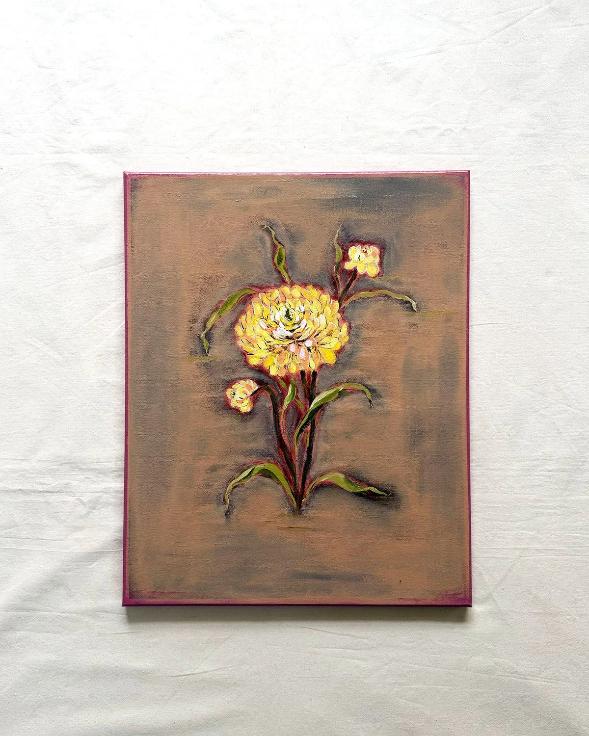 A square canvas painting featuring a stylized yellow flower with green leaves on a textured brown background, bordered in magenta and displayed against a white fabric backdrop.