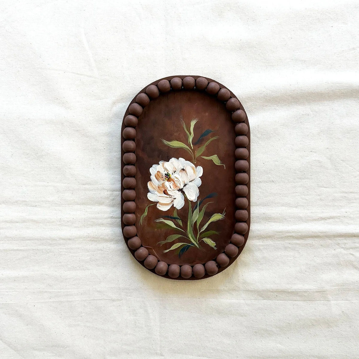 An oval decorative tray with a dark brown beaded border and a hand-painted floral design featuring white flowers and green leaves 
