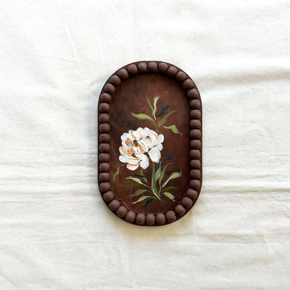An oval decorative tray with a dark brown beaded border and a hand-painted floral design featuring white flowers and green leaves 