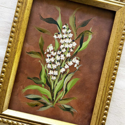 A framed original botanical painting of white lily of the valley flowers with green leaves on a textured brown background, enclosed in an ornate gold frame.