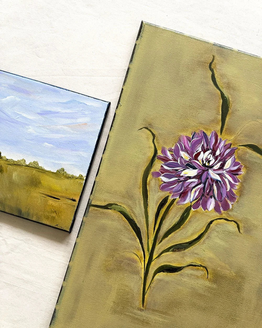 Two small paintings displayed on a light surface: one depicts a purple flower with green leaves on a gold-toned background, and the other shows a landscape with a blue sky and golden field.
