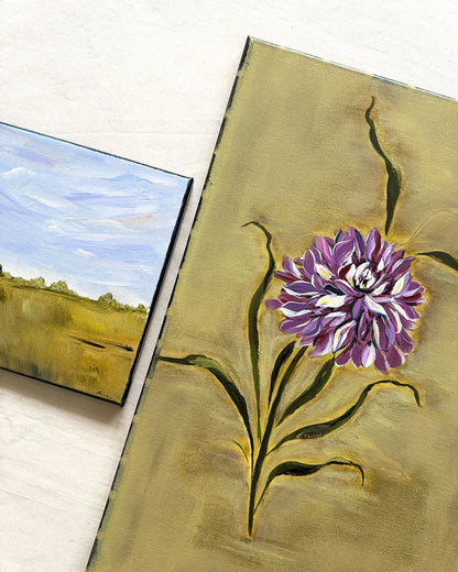 Two small paintings displayed on a light surface: one depicts a purple flower with green leaves on a gold-toned background, and the other shows a landscape with a blue sky and golden field.