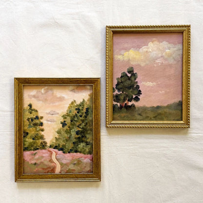 Two framed landscape original paintings displayed side by side. The left painting features a winding path through green trees under a soft pink sky, in a gold frame.