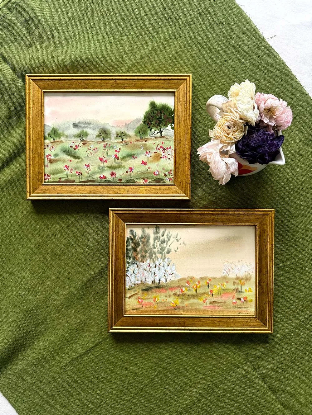 Two small framed landscape paintings with gold frames are arranged on an olive green fabric background, alongside a small vase of pink and purple flowers.