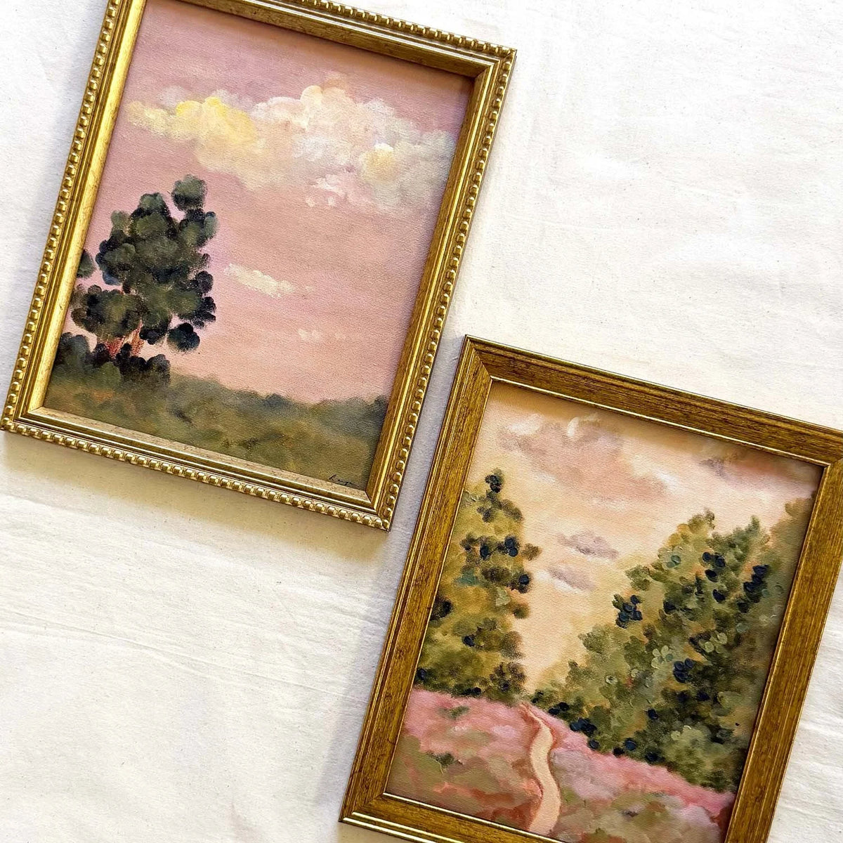 Two framed original landscape paintings with gold frames hanging on a white textured wall. The first painting shows a pink sky with clouds and a dark green tree, while the second painting depicts green trees and a winding pink path under a pale sky.