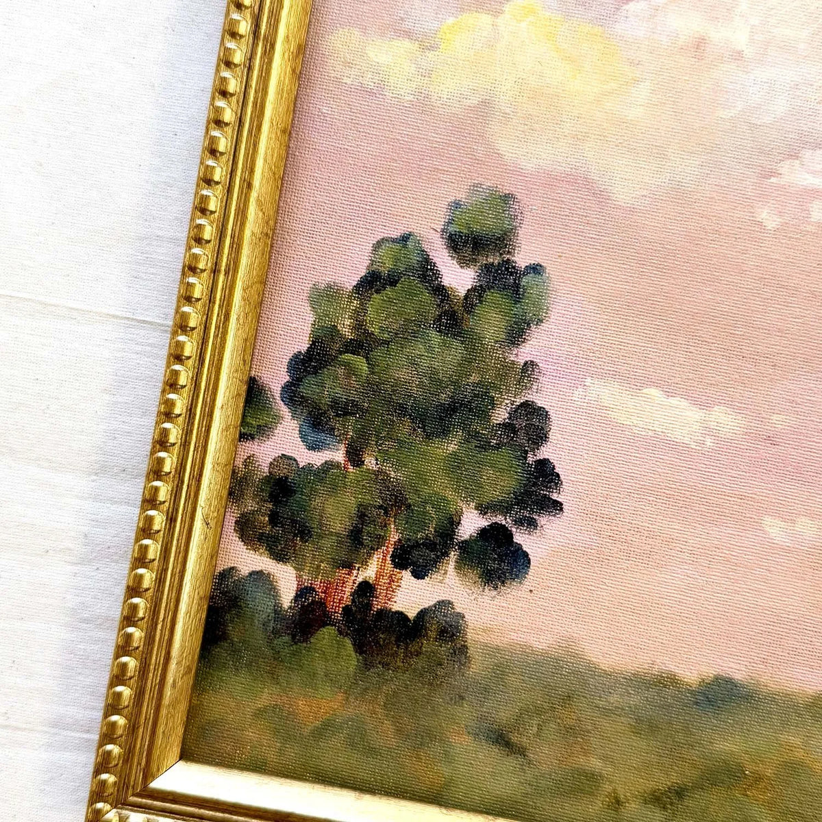 A close-up of a framed landscape painting showing a dark green tree against a soft pink sky with light clouds, with a gold beaded frame visible on the left side.