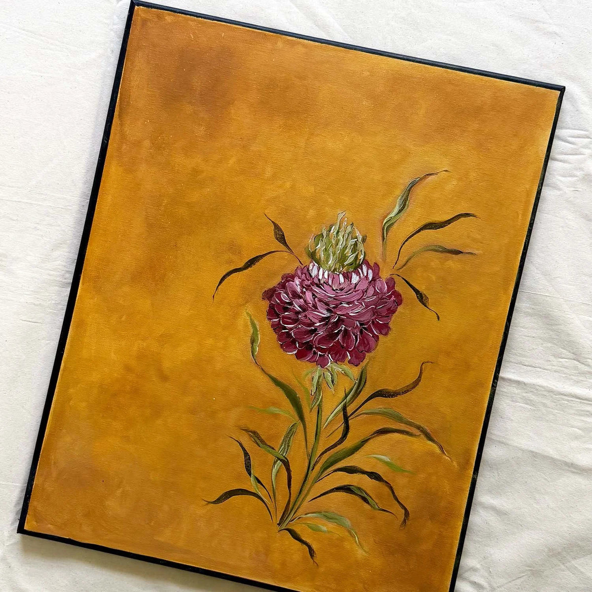 A framed original painting of a deep red ranunculus with green leaves on a textured golden-yellow background