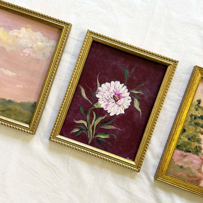 A framed floral painting featuring a white daisy with green leaves on a deep burgundy background, set in a decorative gold frame.