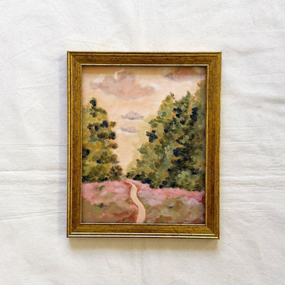 A framed original landscape painting featuring green trees on either side of a winding pink path under a soft beige sky, enclosed in a gold frame 