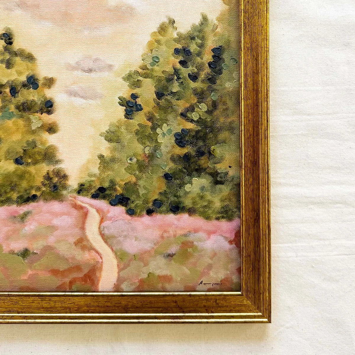 A close-up of a framed landscape original painting showing green trees, a pale pink field, and a winding path, with a gold-toned wooden frame 