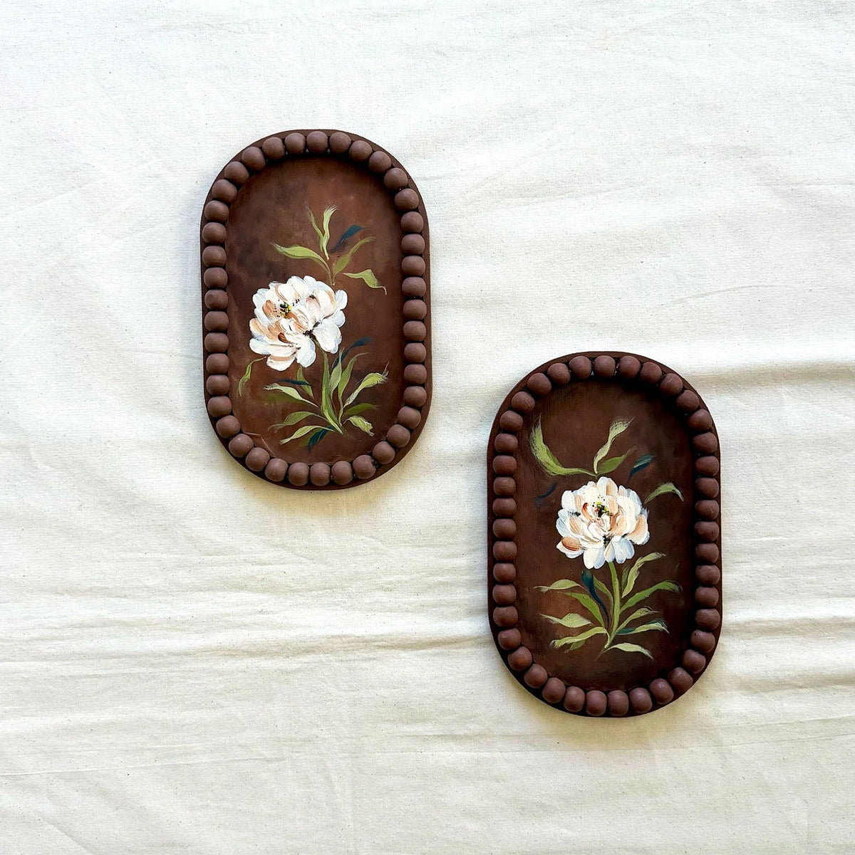 Two oval dark brown trays with beaded borders and painted white flowers with green leaves, displayed on a white fabric background.