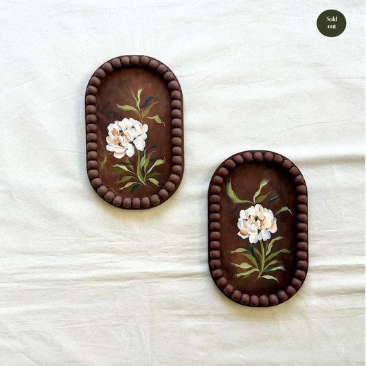 Two oval dark brown trays with beaded borders and hand-painted white flowers with green leaves, displayed on a textured off-white fabric background.