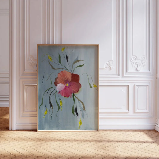 A framed botanical art print featuring a pink and red flower with green leaves on a muted blue-gray background, leaning against a white paneled wall on a light wood herringbone floor.