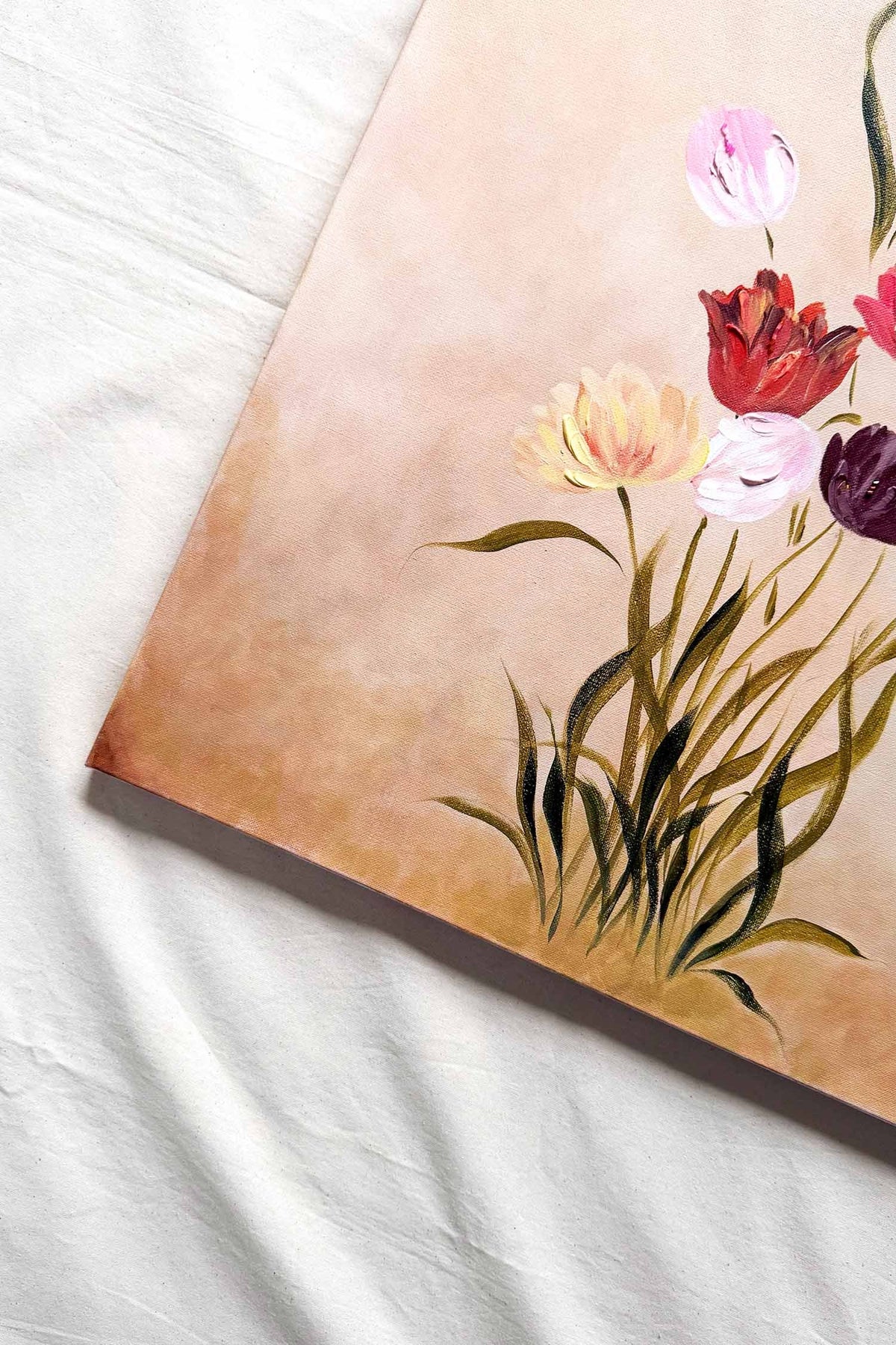 A close-up of a floral original painting on a canvas, showing tulips in pink, red, pale yellow, and purple with green leaves on a warm beige background.