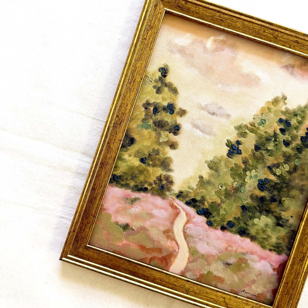 A framed original landscape painting featuring green trees, a pinkish path, and a soft beige sky, enclosed in a gold-toned frame. Original by Ramya Rajiv